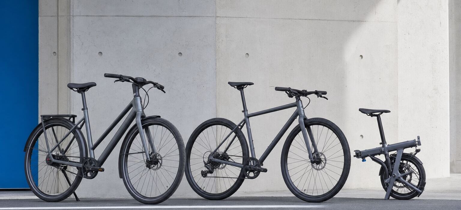 Tre biciclette da città moderne accostate a una parete: una commuter con parafanghi e trasmissione a cinghia, una ibrida flat-bar e una pieghevole